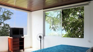 A bedroom with a large window, a bed with a blue quilt, a wooden dresser, and a television on top. - Finca Hotel Villa Gladys (El Colegio)