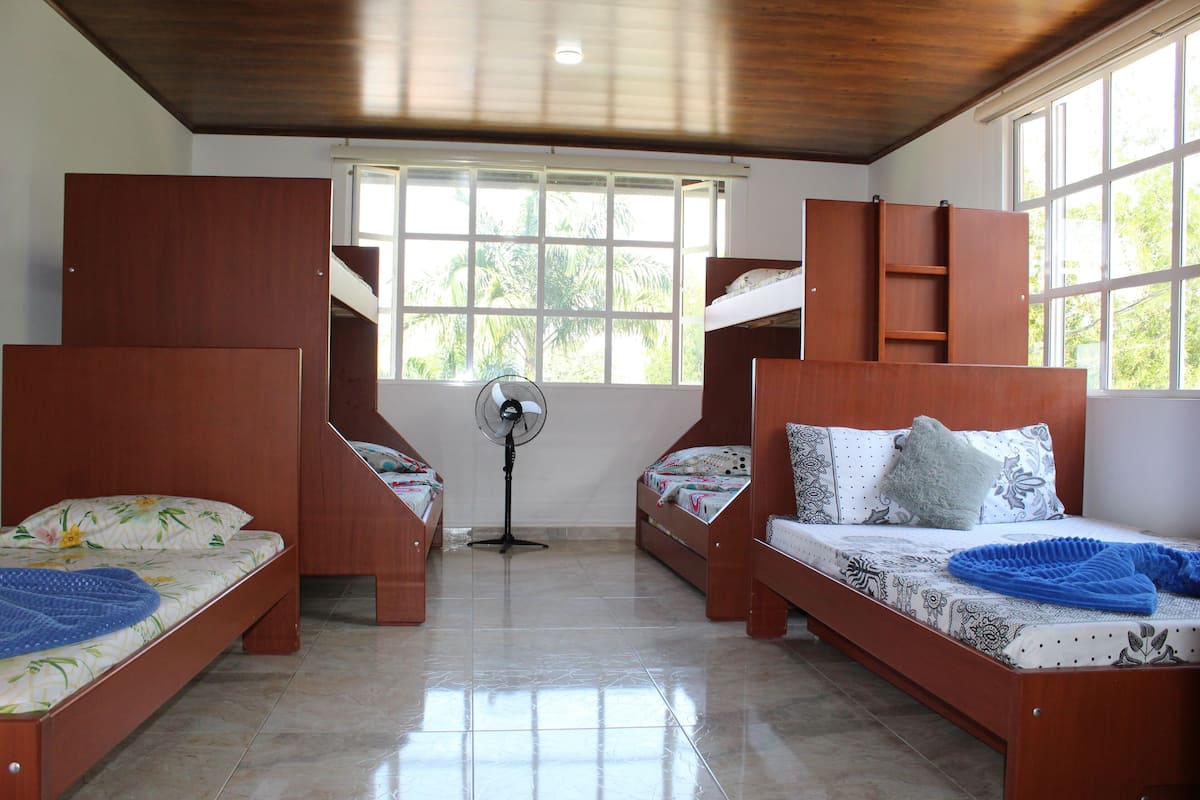 A room with bunk beds, a standing fan, and large windows.
