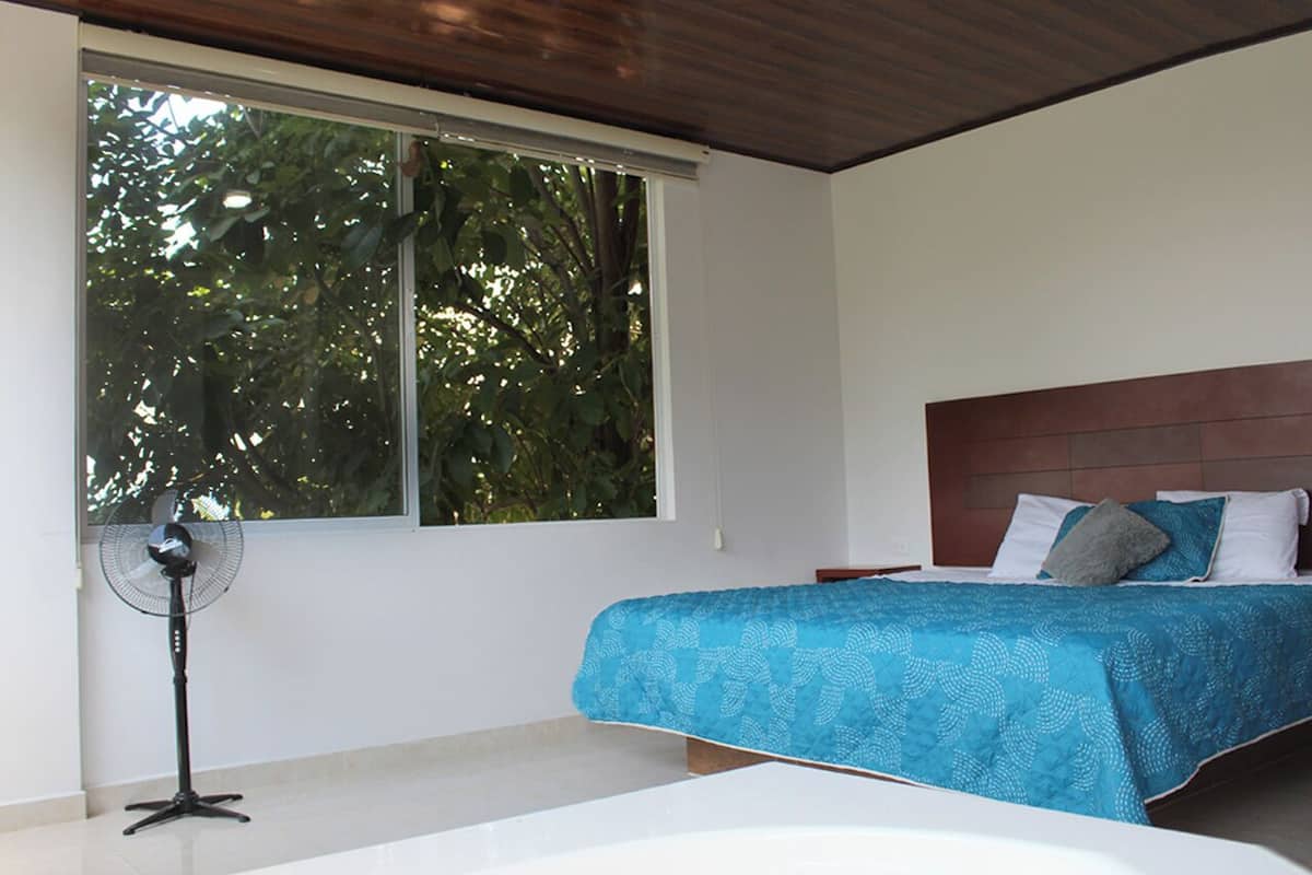 A bedroom with a bed, a standing fan, and a large window with a view of trees.
