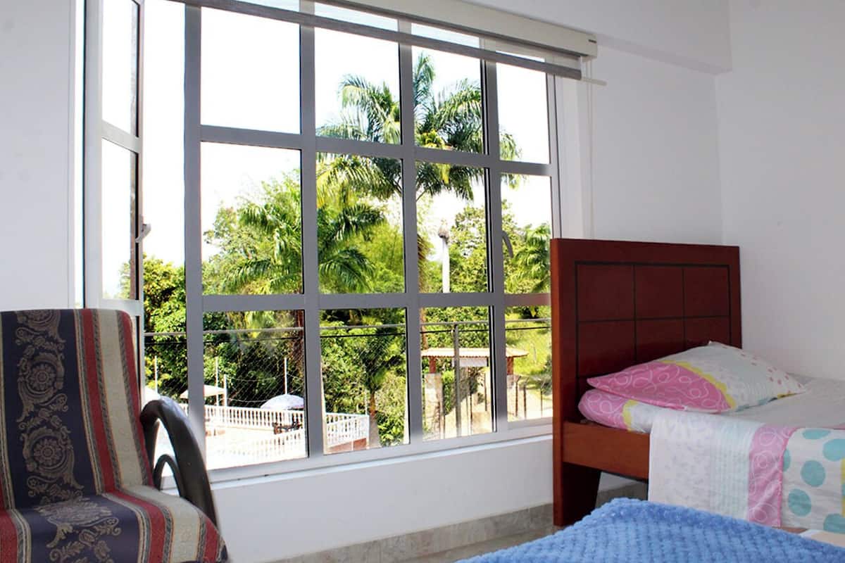 A room with a large window, a bed, a chair, and a view of palm trees outside.