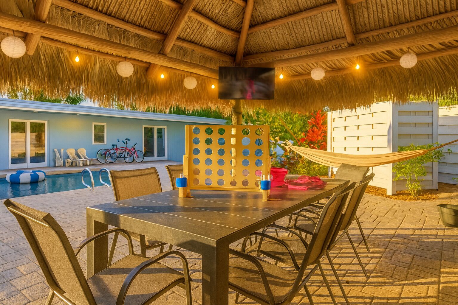 Un área de comedor al aire libre con mesa y sillas, una piscina y una estructura con techo de paja.