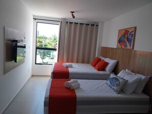 Two single beds with white bedding and red accents, a flat-screen TV, and a large window with beige curtains.