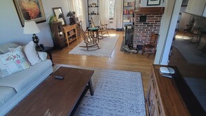 A spacious living room with a sofa, coffee table, and rocking chairs. There is a fireplace with a brick wall and a bookshelf filled with books. - Experience Classic Charm in a Cozy 115-Year-Old Colonial (Waltham)