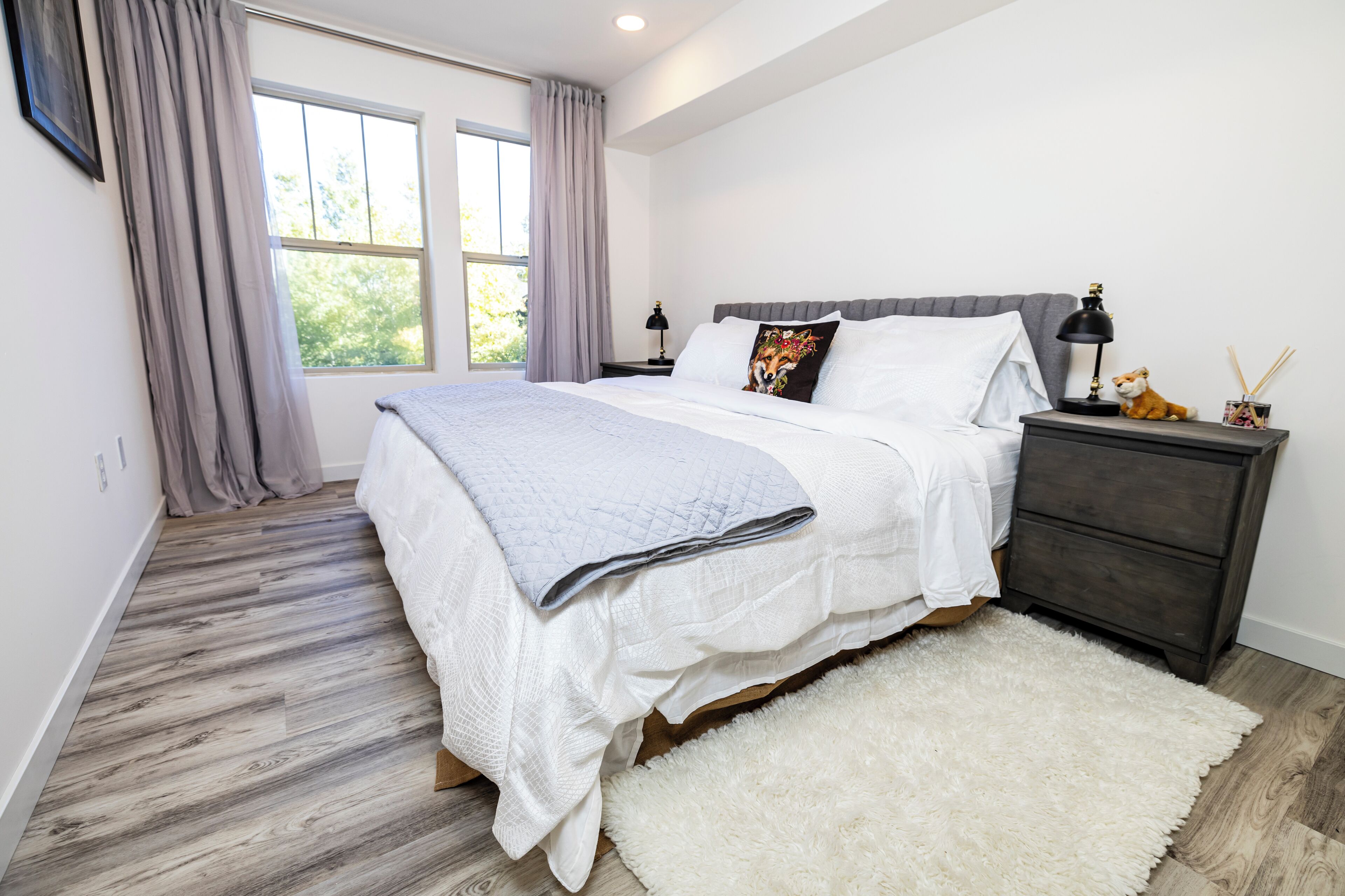 A bedroom with a large bed, a nightstand with a lamp, and a view of the outdoors through a window.