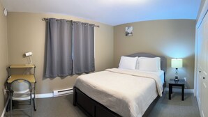 Hotellihuoneessa on sänky, työpöytä tuolilla, yöpöytä lampun kanssa ja taulu seinällä.