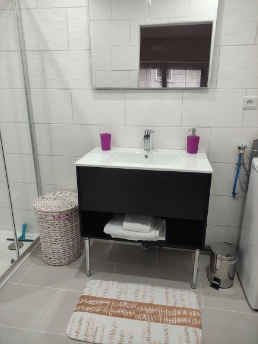 A bathroom with a white sink, two purple cups, a mirror, a wicker basket, a white towel, and a patterned rug.