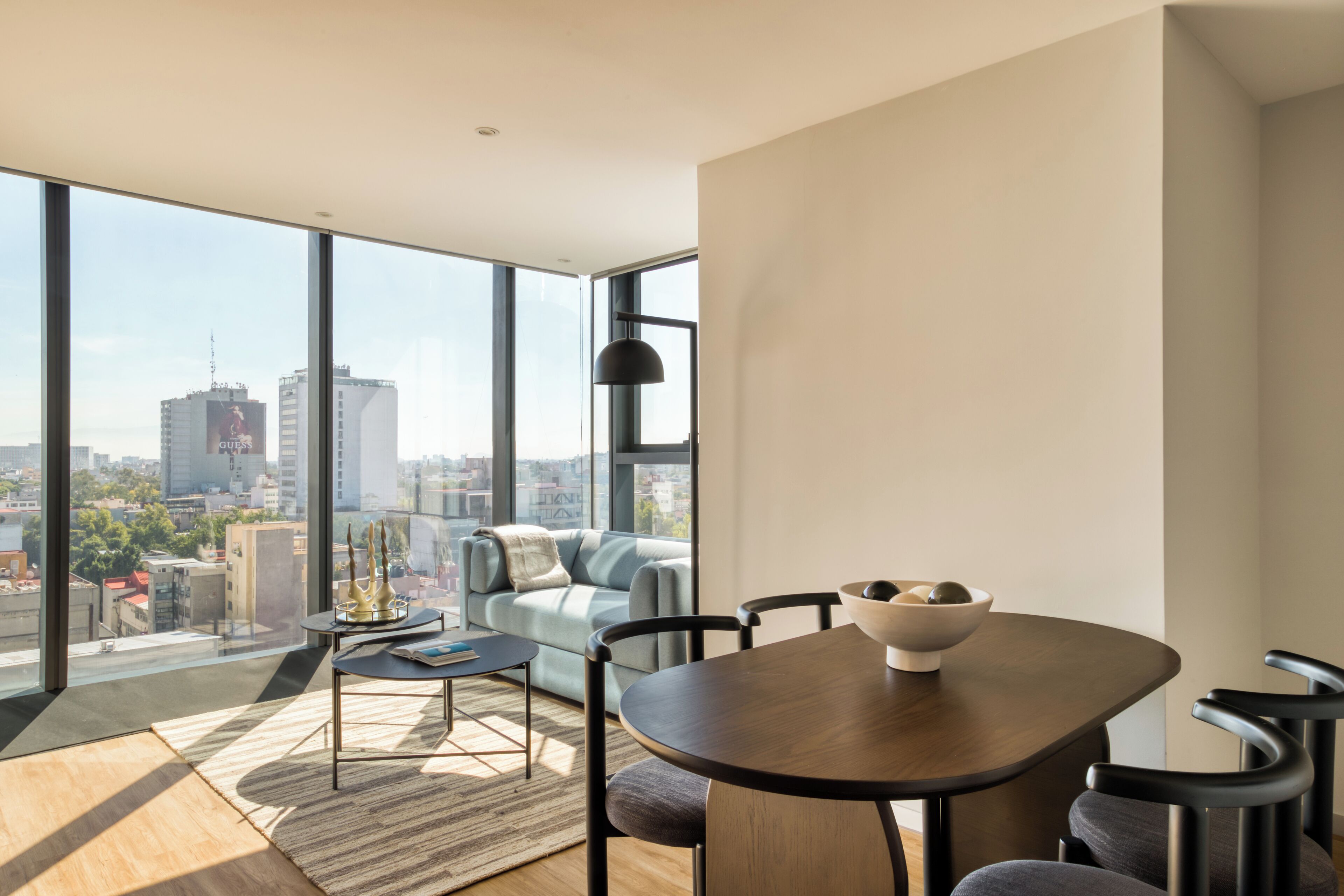 A modern living room with a dining table, a sofa, and a large window offering a city view.