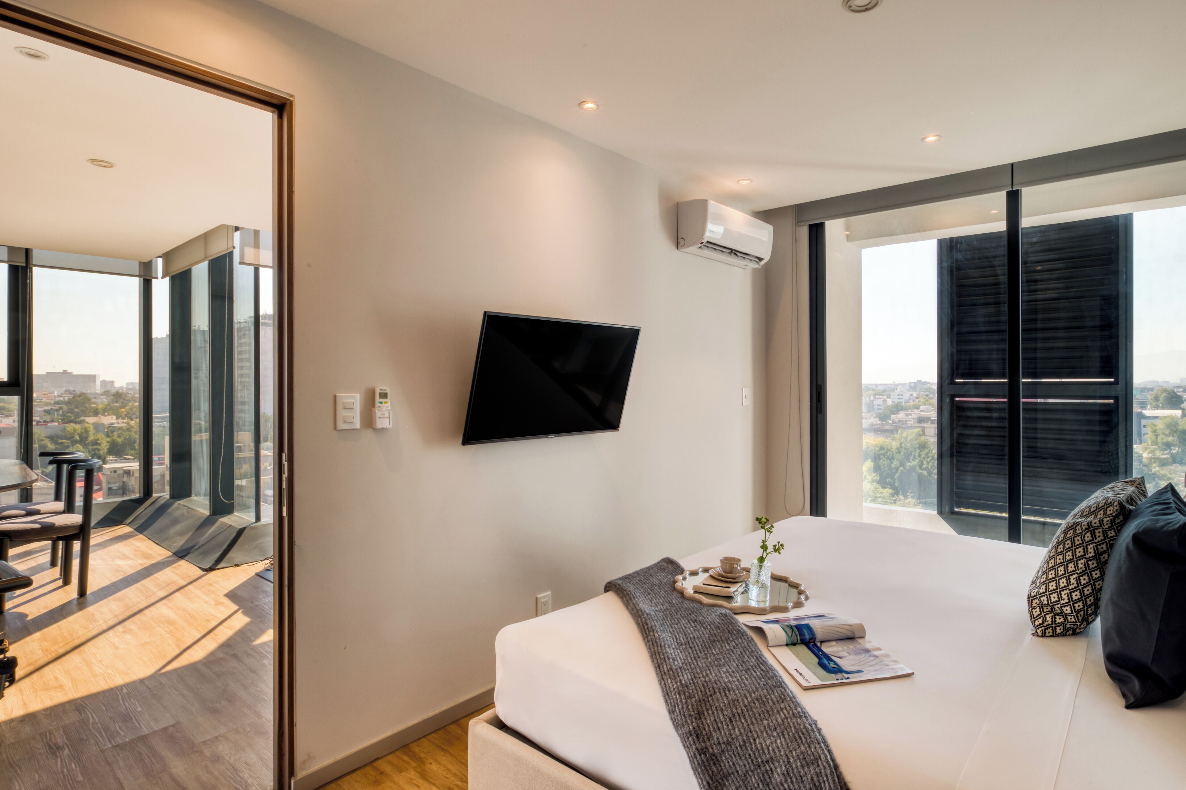 A modern bedroom with a large bed, a flat-screen TV mounted on the wall, and a city view through large windows.
