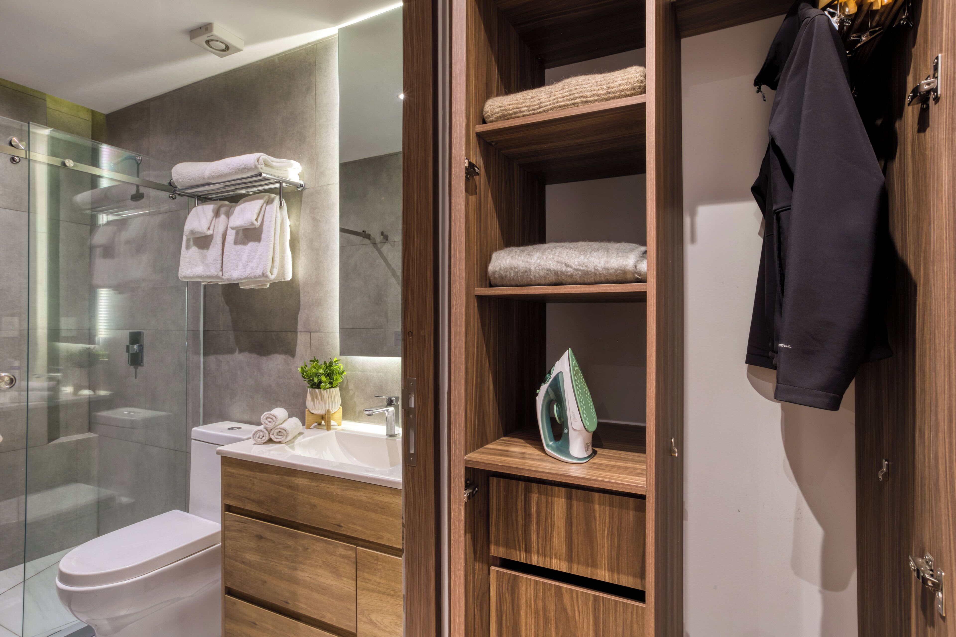 A modern bathroom with a glass shower enclosure, a wooden vanity with a sink, and a built-in shelving unit with towels and an iron.