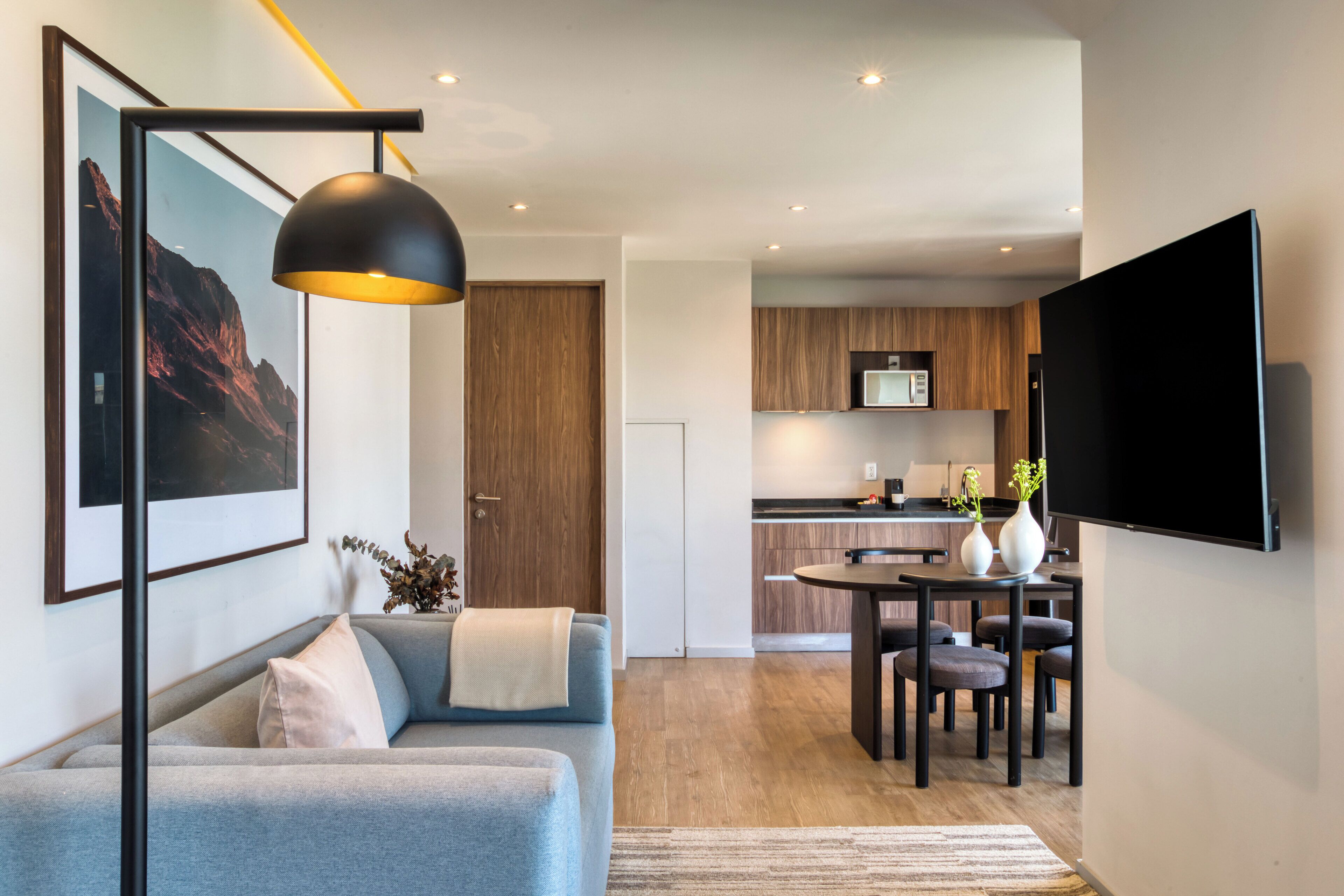 A modern living room with a sofa, a dining table with chairs, a TV mounted on the wall, and a kitchen in the background.