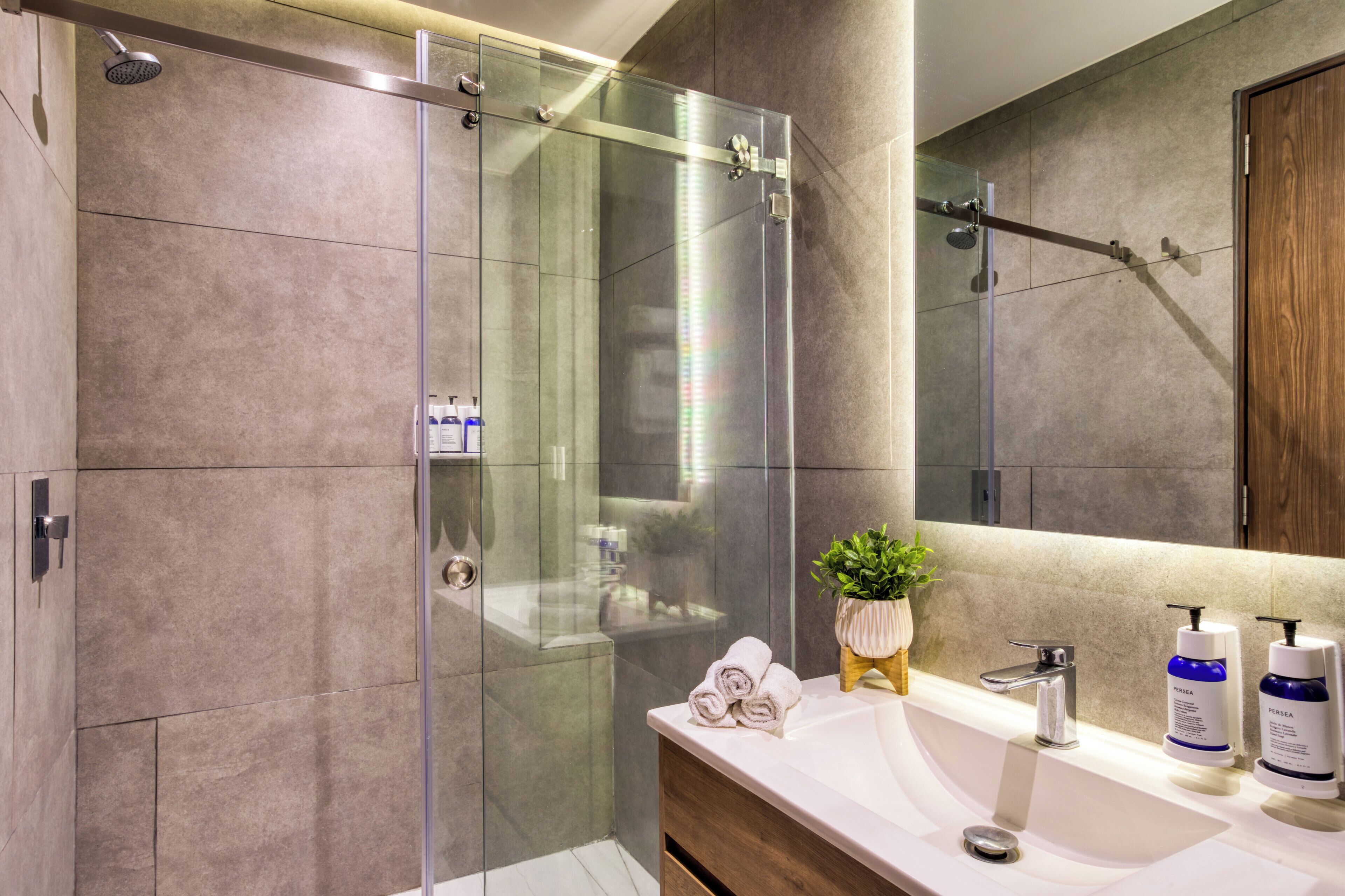 A modern bathroom with a glass-enclosed shower, a white sink with two bottles of shampoo, and a wooden cabinet.