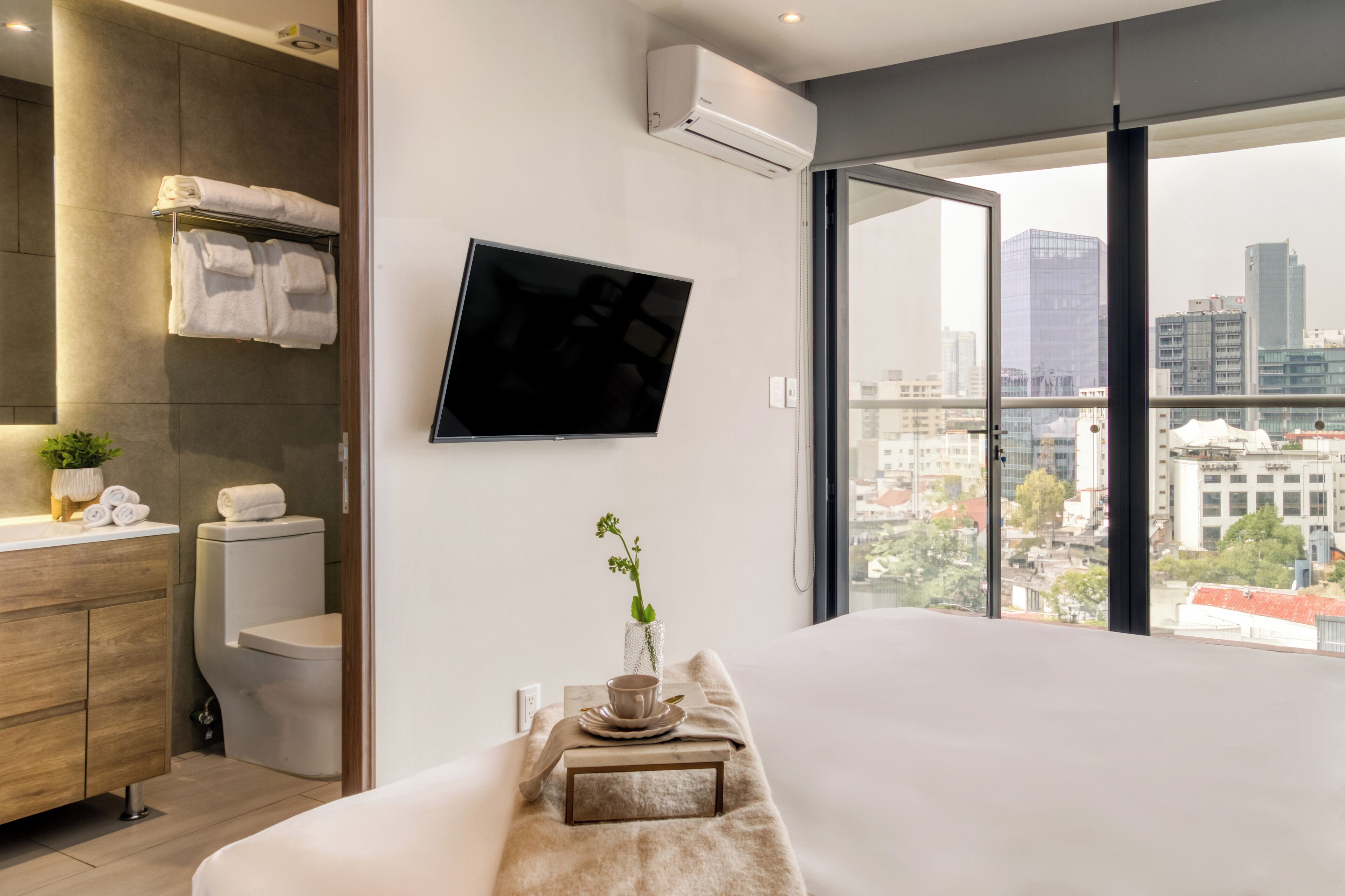 Camera d'albergo moderna con un ampio letto, una TV a schermo piatto montata a parete, vista sulla città attraverso ampie finestre, una toeletta in legno con specchio e una piccola pianta in vaso.