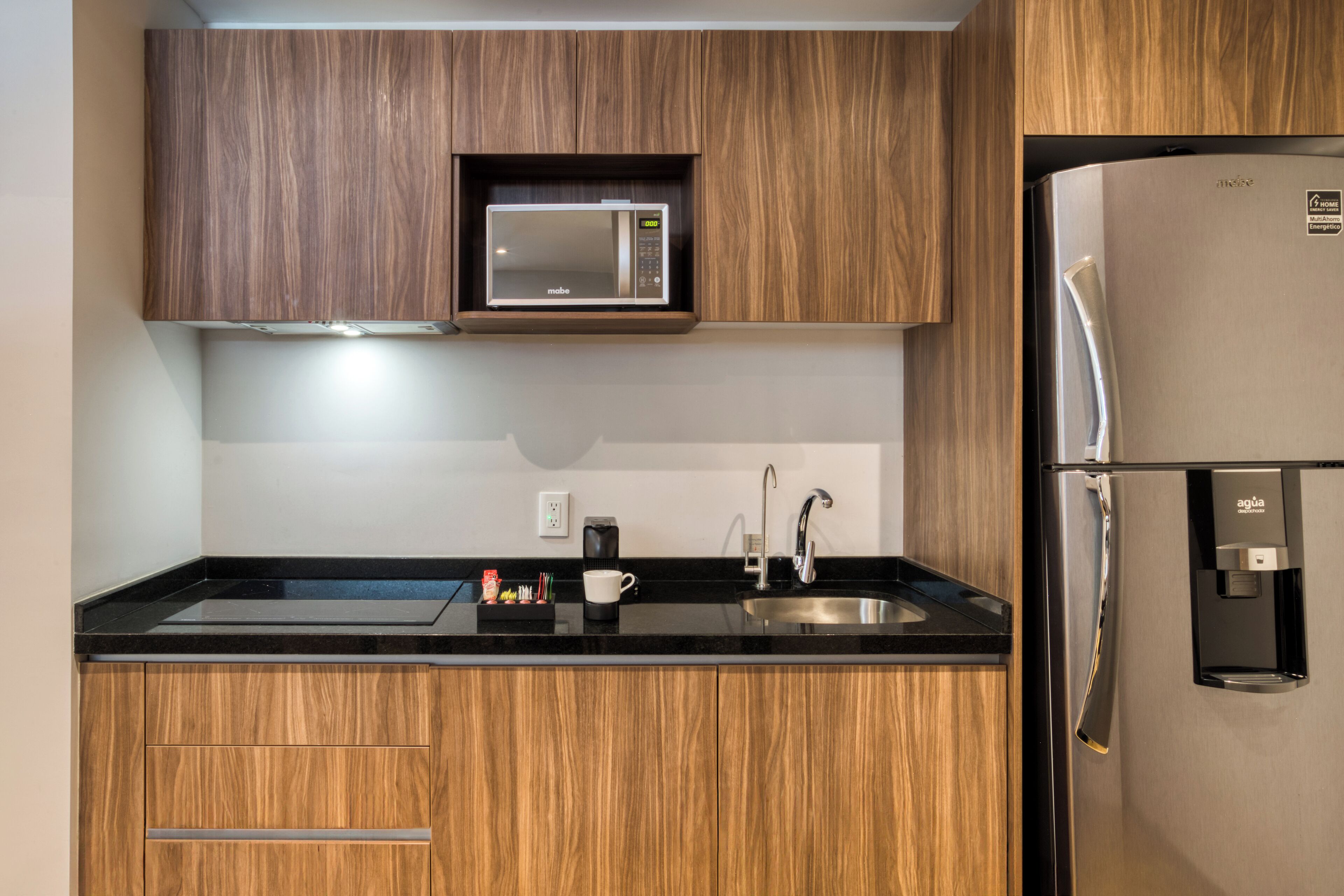 A modern kitchen with wooden cabinets, a built-in microwave, a stainless steel refrigerator, and a black countertop.