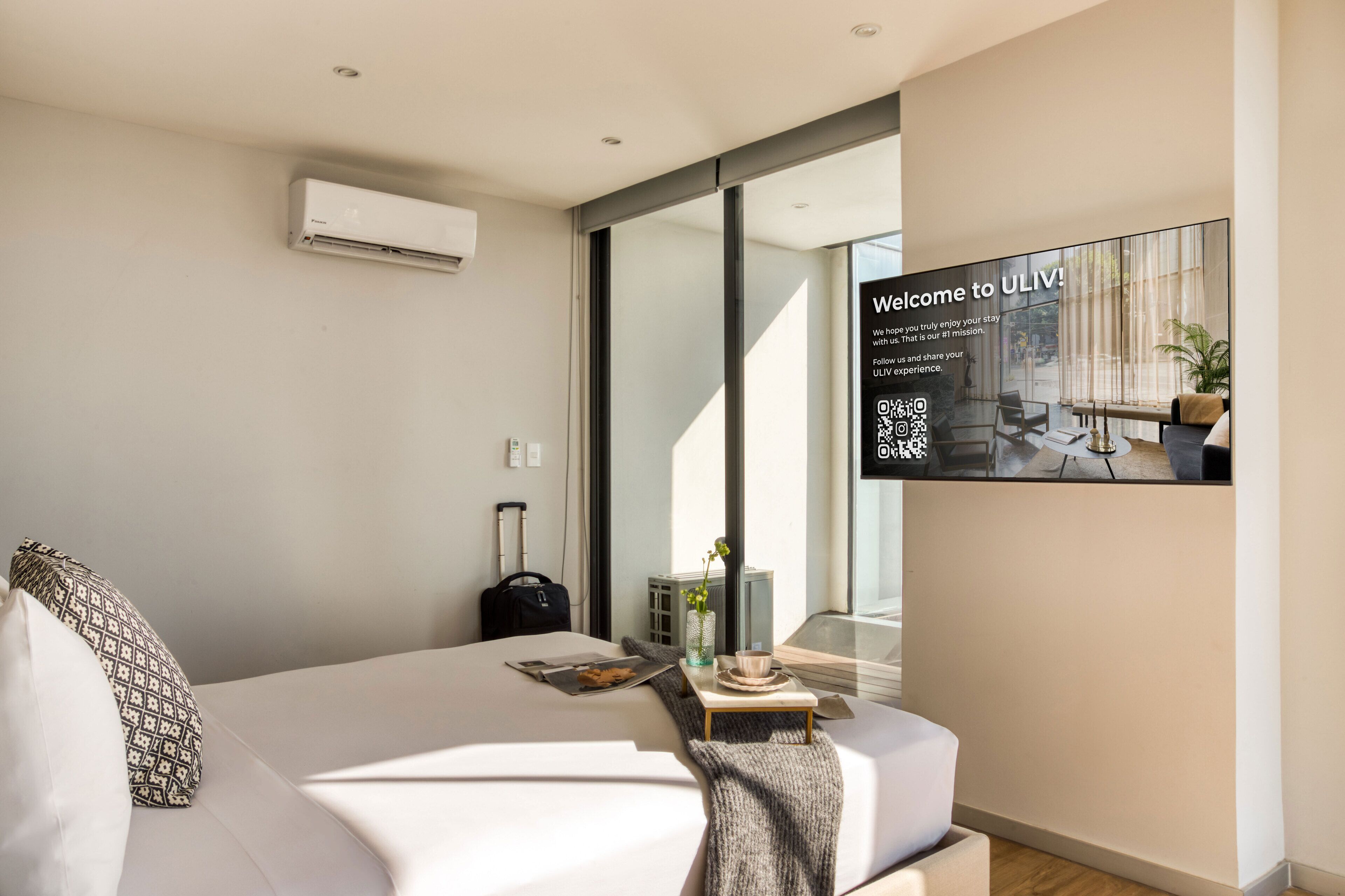 Una habitación de hotel moderna con una cama grande, una mesita con una taza y un platillo, una maleta y un aire acondicionado instalado en la pared.