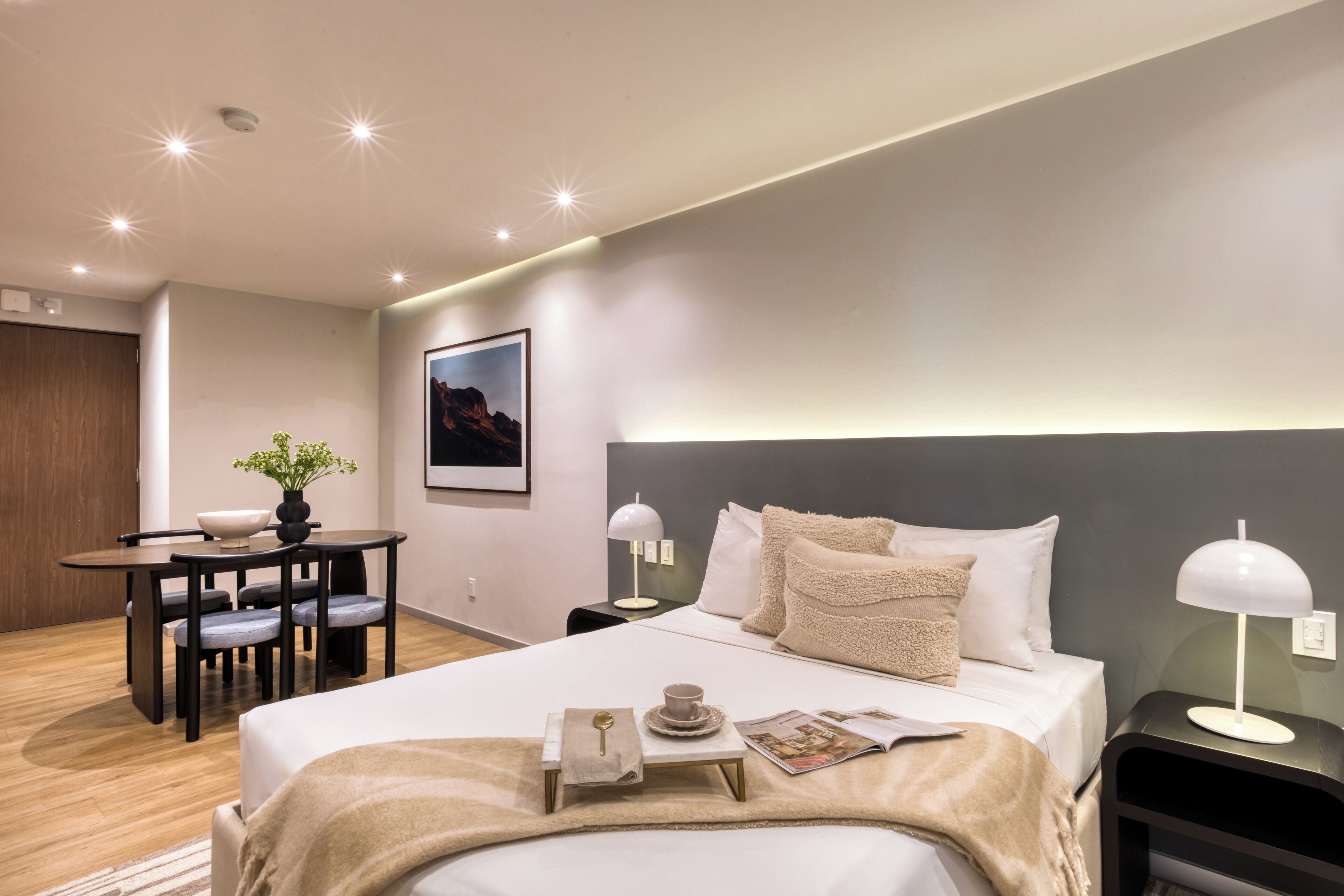 Una habitación de hotel moderna con una cama grande, mesitas de noche con lámparas, un escritorio pequeño con una silla y una planta en maceta.