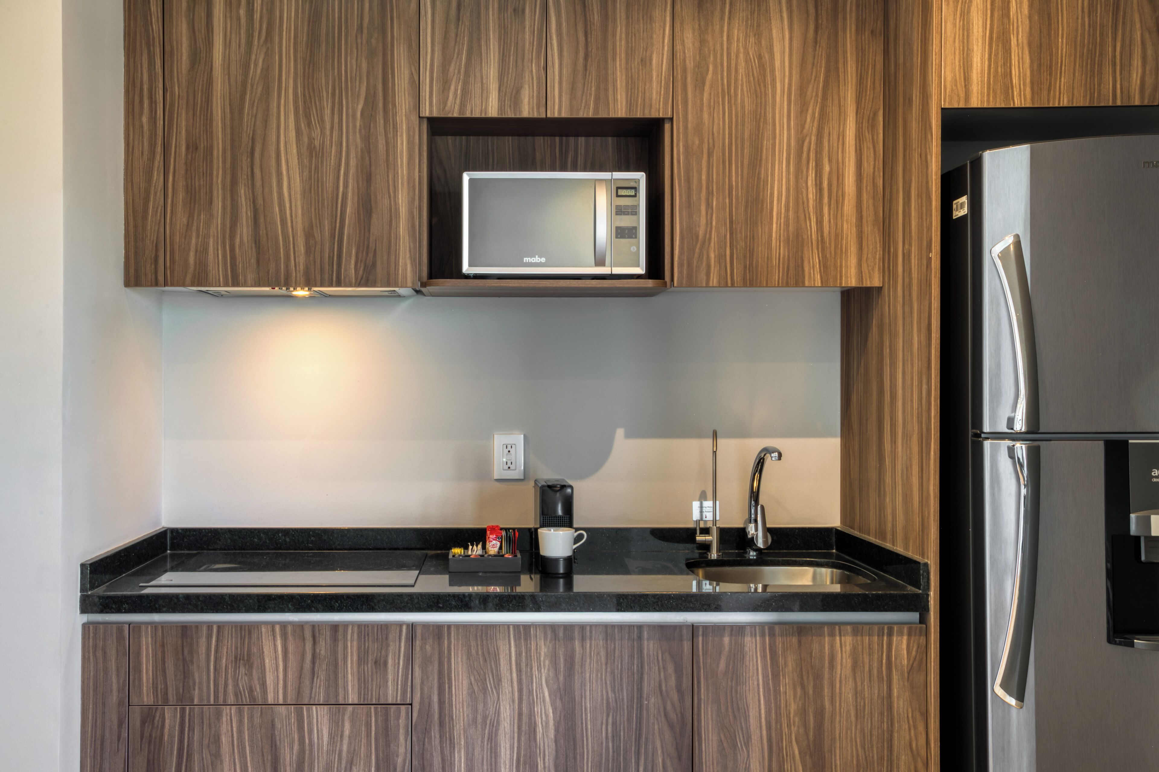 A modern kitchen with wooden cabinets, a built-in microwave, a stainless steel refrigerator, a coffee maker, and a sink.