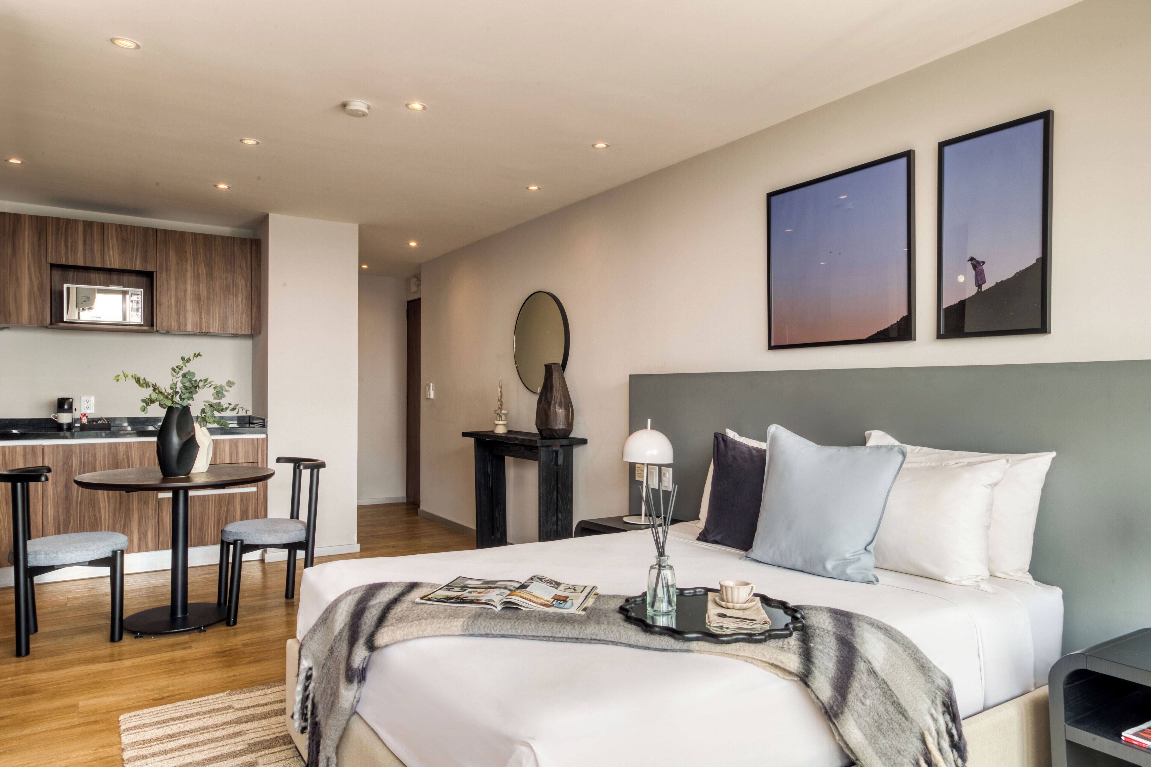 A modern hotel room with a large bed, a dining area with a table and chairs, a kitchen in the background, and two framed pictures on the wall.