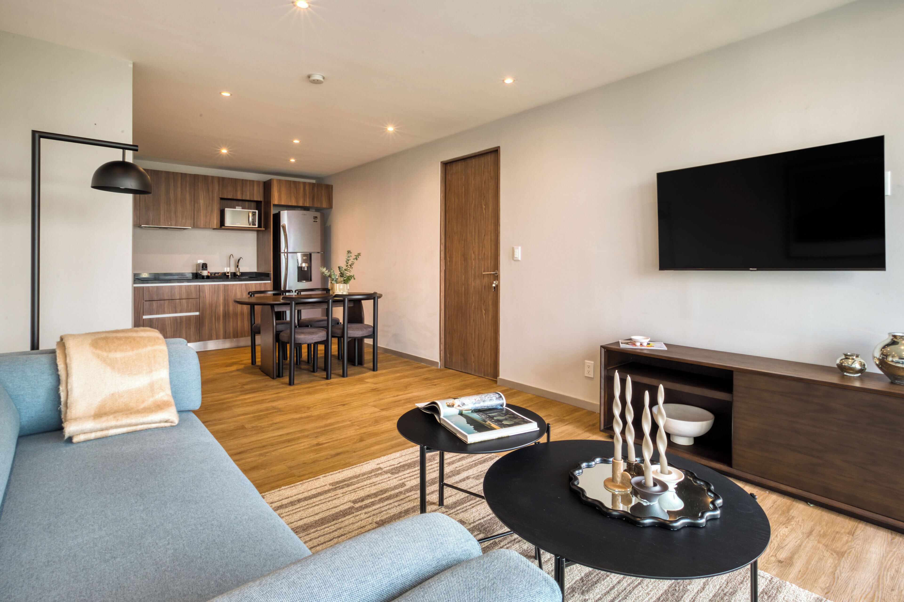 Una sala de estar moderna con un sofá, una mesa de centro y una zona de comedor con una mesa y sillas. Se ve una cocina al fondo con un televisor de pantalla plana montado en la pared.