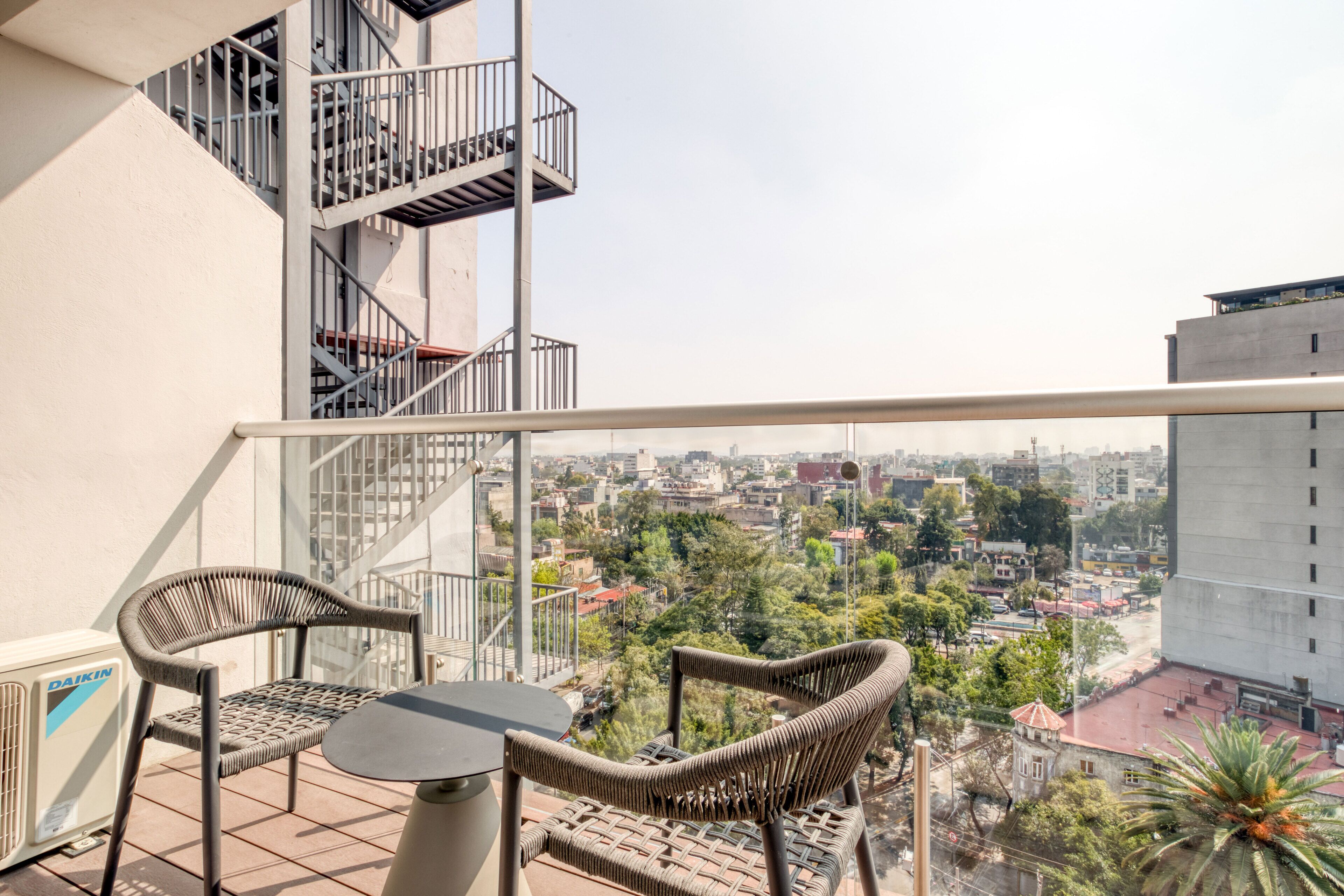 A balcony with wicker chairs and a table, overlooking a cityscape with buildings and trees.