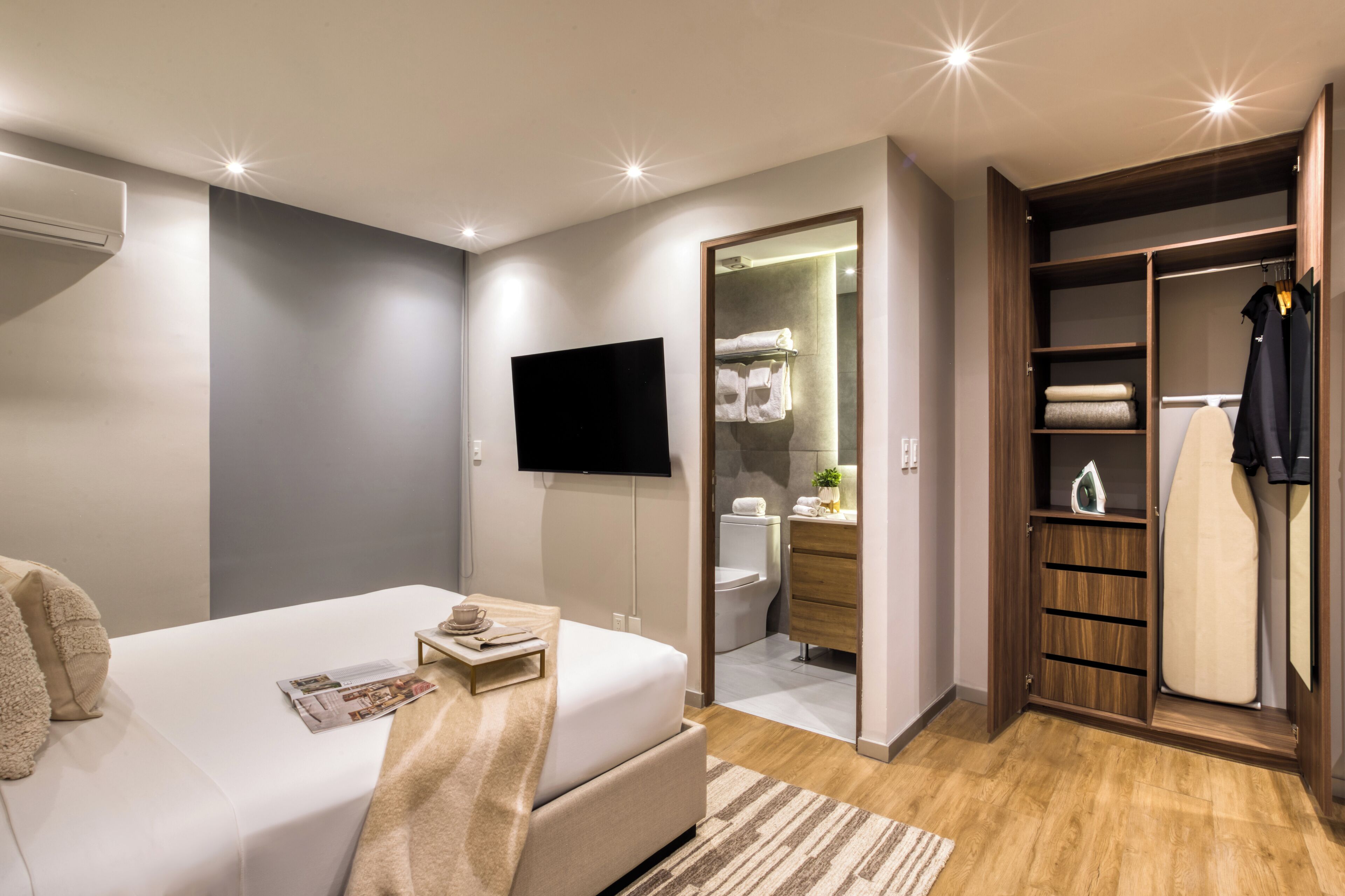 Een moderne slaapkamer met een bed, nachtkastje, televisie en een inloopkast met spiegel en hangende kleding.