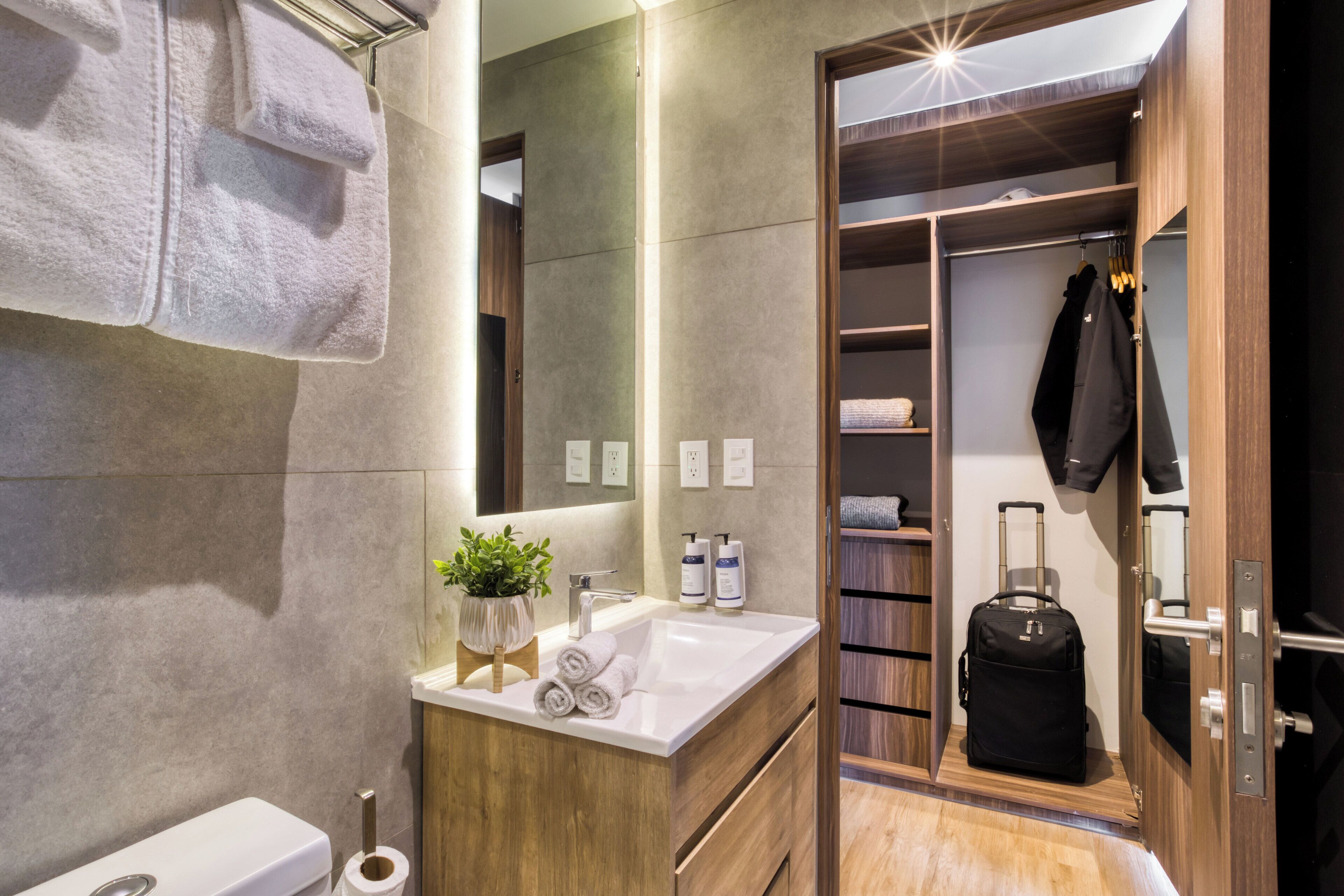 A modern bathroom with a wooden vanity, a sink, and a mirror. There is a towel rack with towels, a potted plant, and toiletries on the vanity. A suitcase is visible in the closet.