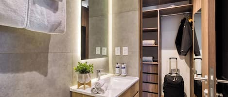 A modern bathroom with a wooden vanity, a sink, and a mirror. There is a towel rack with towels, a potted plant, and toiletries on the vanity. A suitcase is visible in the closet.