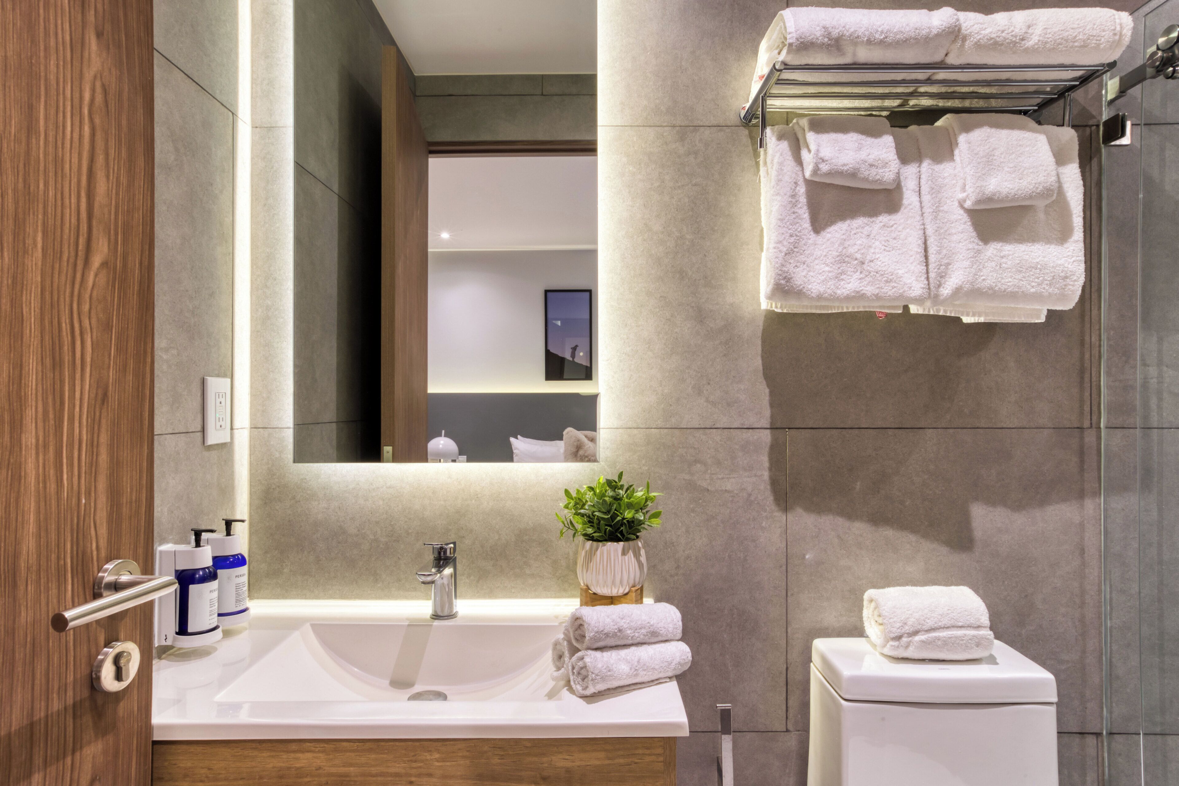 Une salle de bain moderne avec un grand lavabo, un miroir et des toilettes. Des serviettes sont suspendues sur un porte-serviettes et une plante en pot est posée sur le lavabo.