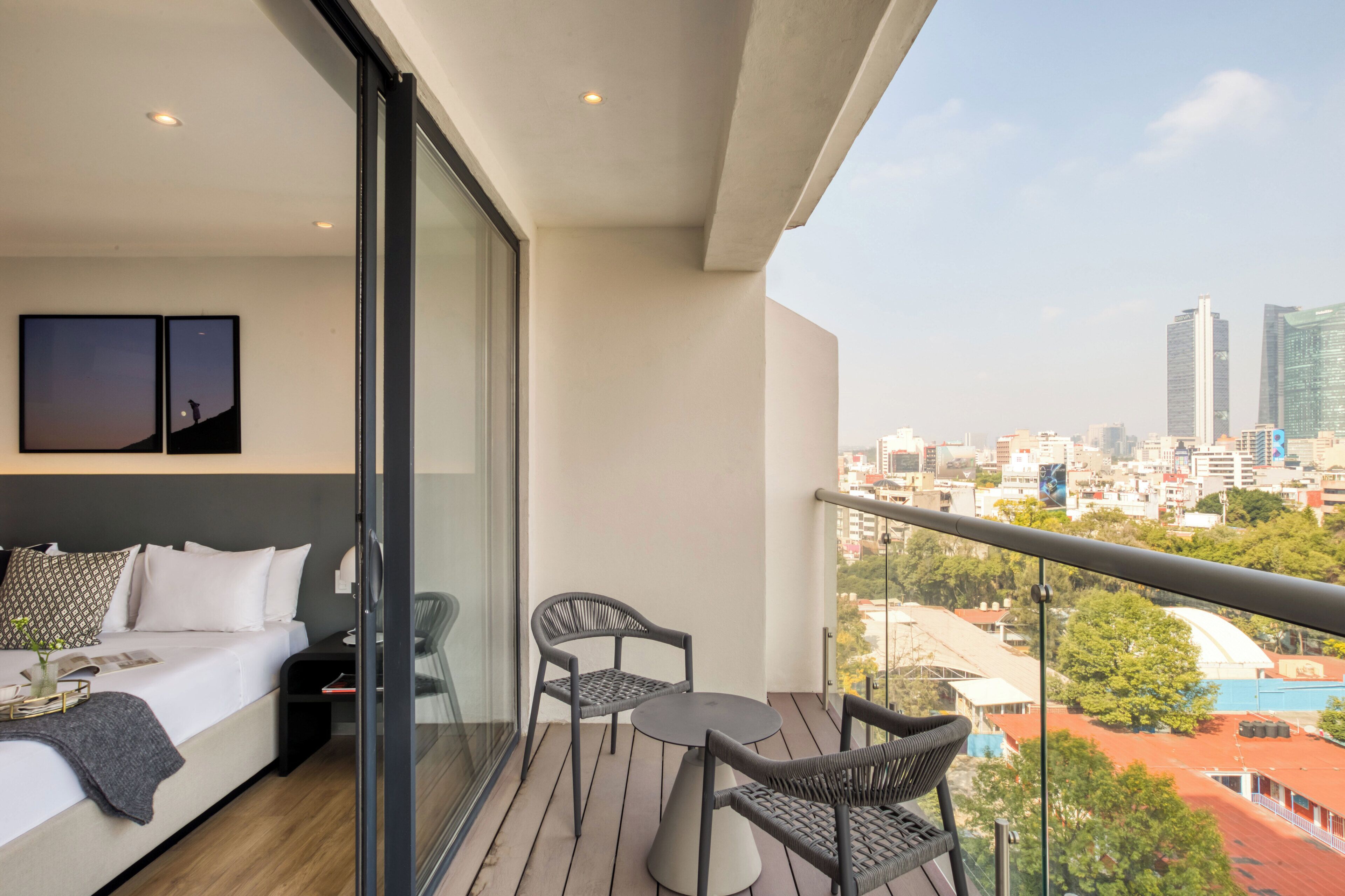 A modern hotel room with a balcony, a bed with white linens, two wicker chairs, and a small table.
