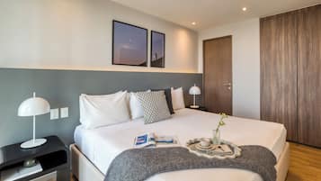 Une chambre d’hôtel moderne avec un lit soigneusement fait, une table de chevet avec une lampe, un plateau avec une tasse et une plante, et une porte avec un panneau en bois.
