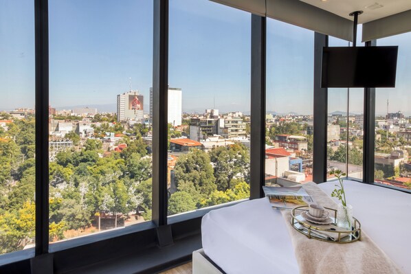 A hotel room with a large window offering a city view, a bed, a bedside table with a tray, and a flat-screen TV. - ULIV Mexico City (Mexico City)