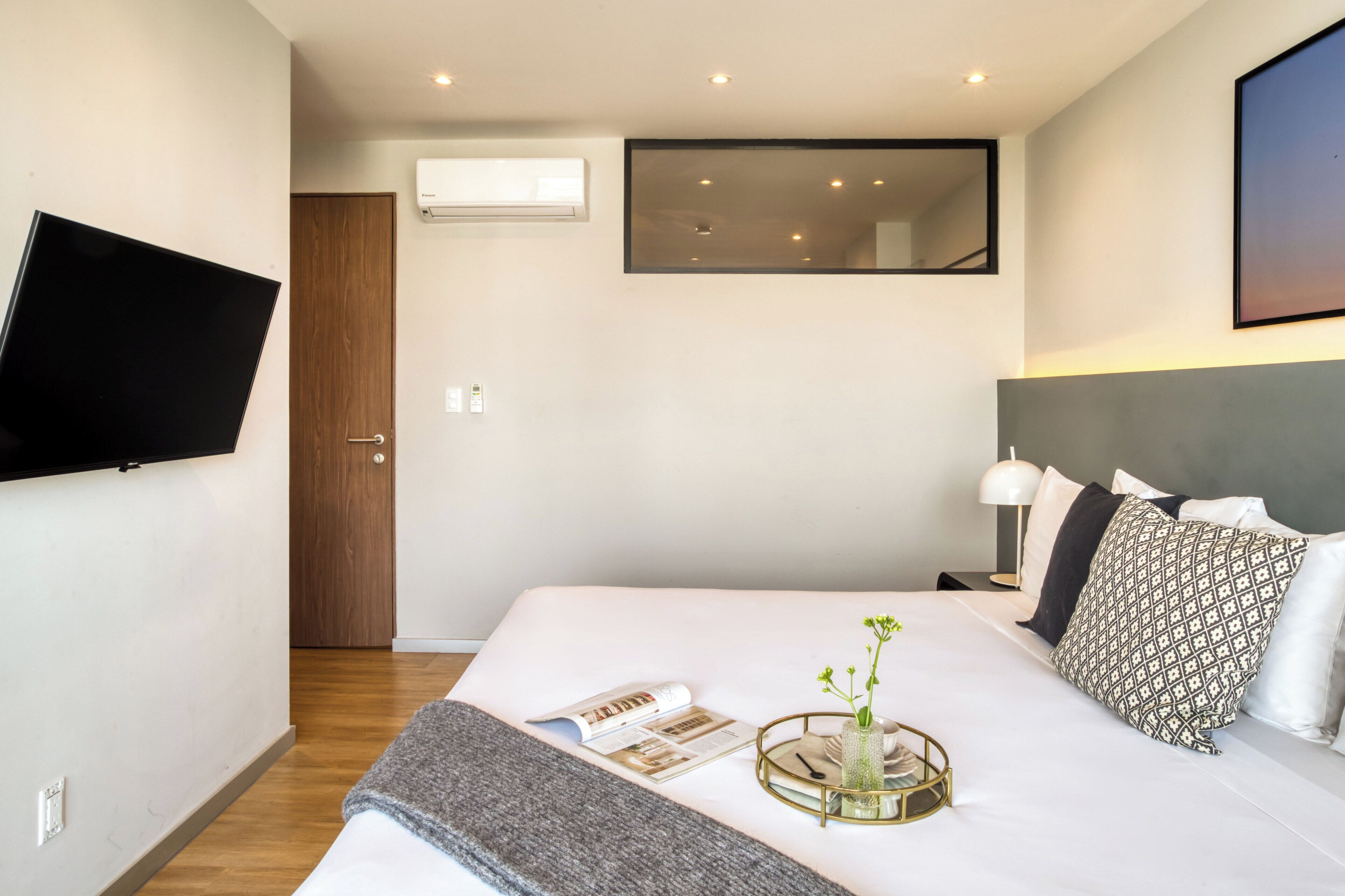 Una habitación de hotel moderna con una televisión de pantalla plana montada en la pared, una cama grande con cabecero, una mesita de noche con lámpara y una ventana con vistas al cielo.
