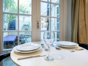 Une table à manger est dressée avec des assiettes, des couverts et un verre à vin, et est placée près d’une fenêtre d’où l’on peut admirer un bâtiment et de la verdure.