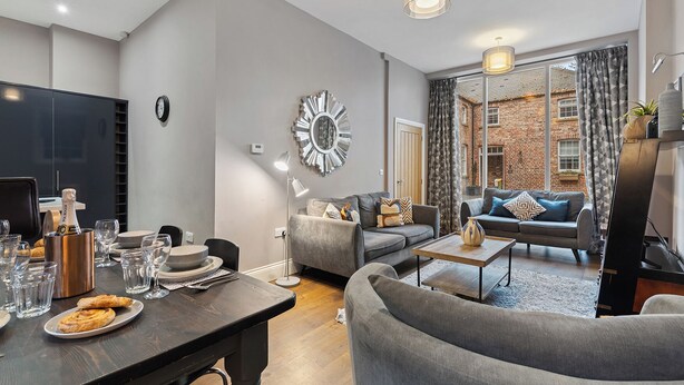 A modern living room with a dining area, grey sofas, a large mirror, and a brick exterior view. - No. 1 - Laurel Cottage - Pass the Keys (York)