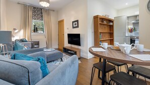 A modern living room with a flat-screen TV, grey sofas, a wooden dining table with chairs, and a kitchen area with cabinets and a microwave. - No. 4 - Oak Tree Cottage - Pass the Keys (Middlethorpe)