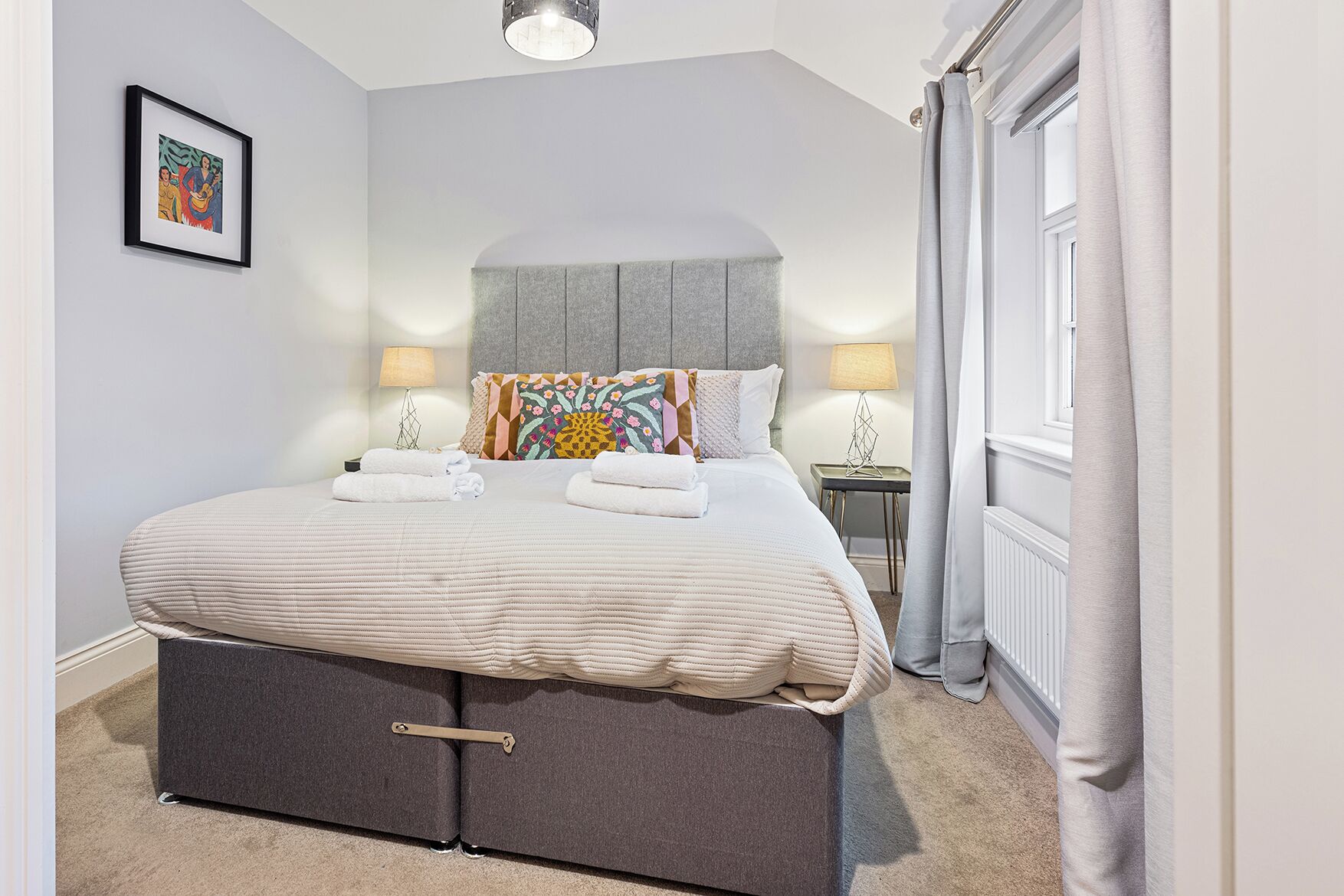 A neatly made bed with a grey headboard, two bedside lamps, and a framed picture on the wall.