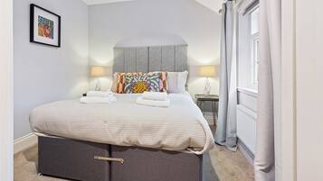 A neatly made bed with a grey headboard, two bedside lamps, and a framed picture on the wall.