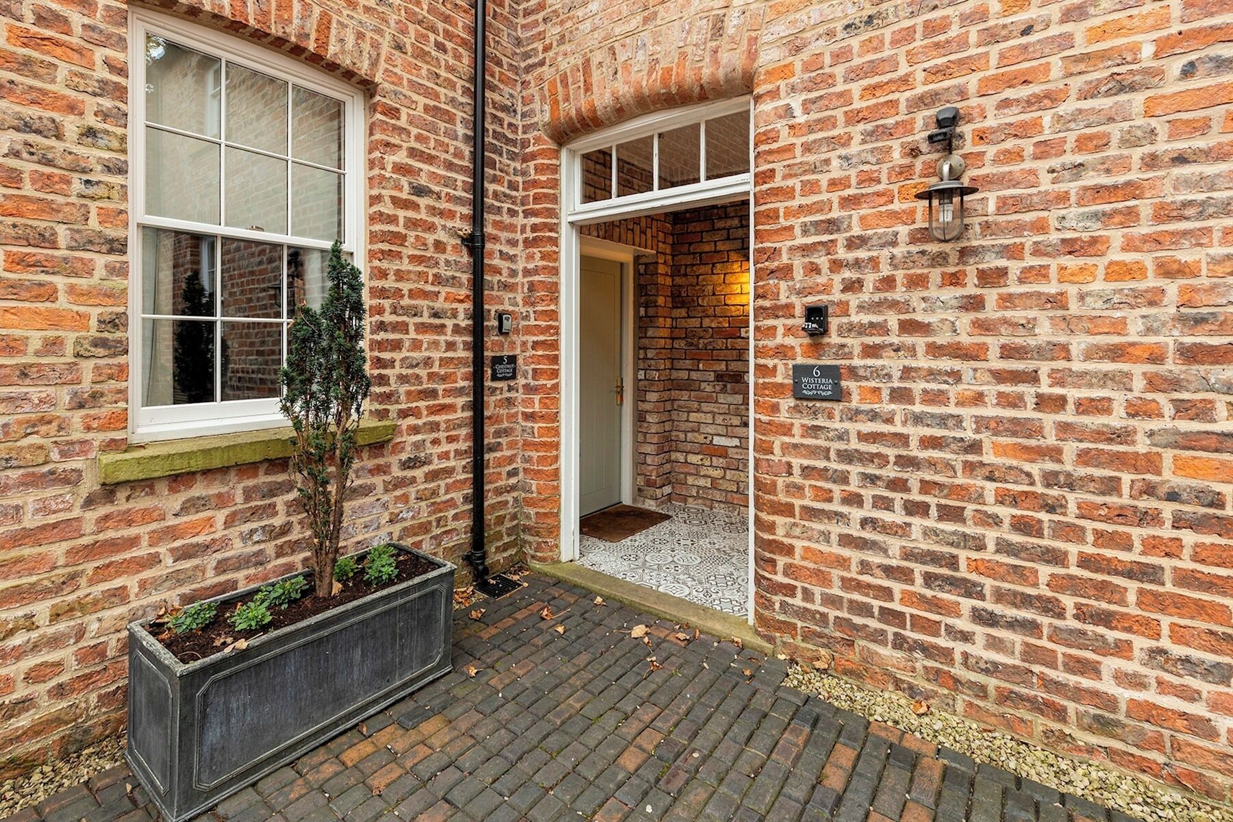 A brick building with two windows and a door, a small planter with plants, and a patterned floor.