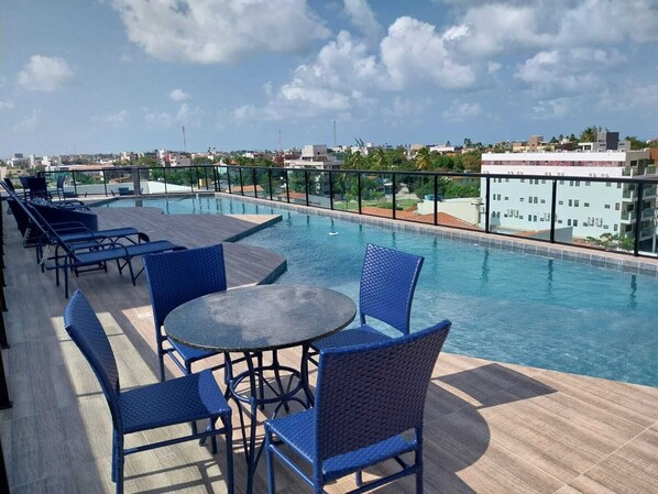 A rooftop pool area with blue chairs and a table, surrounded by buildings and a clear sky.