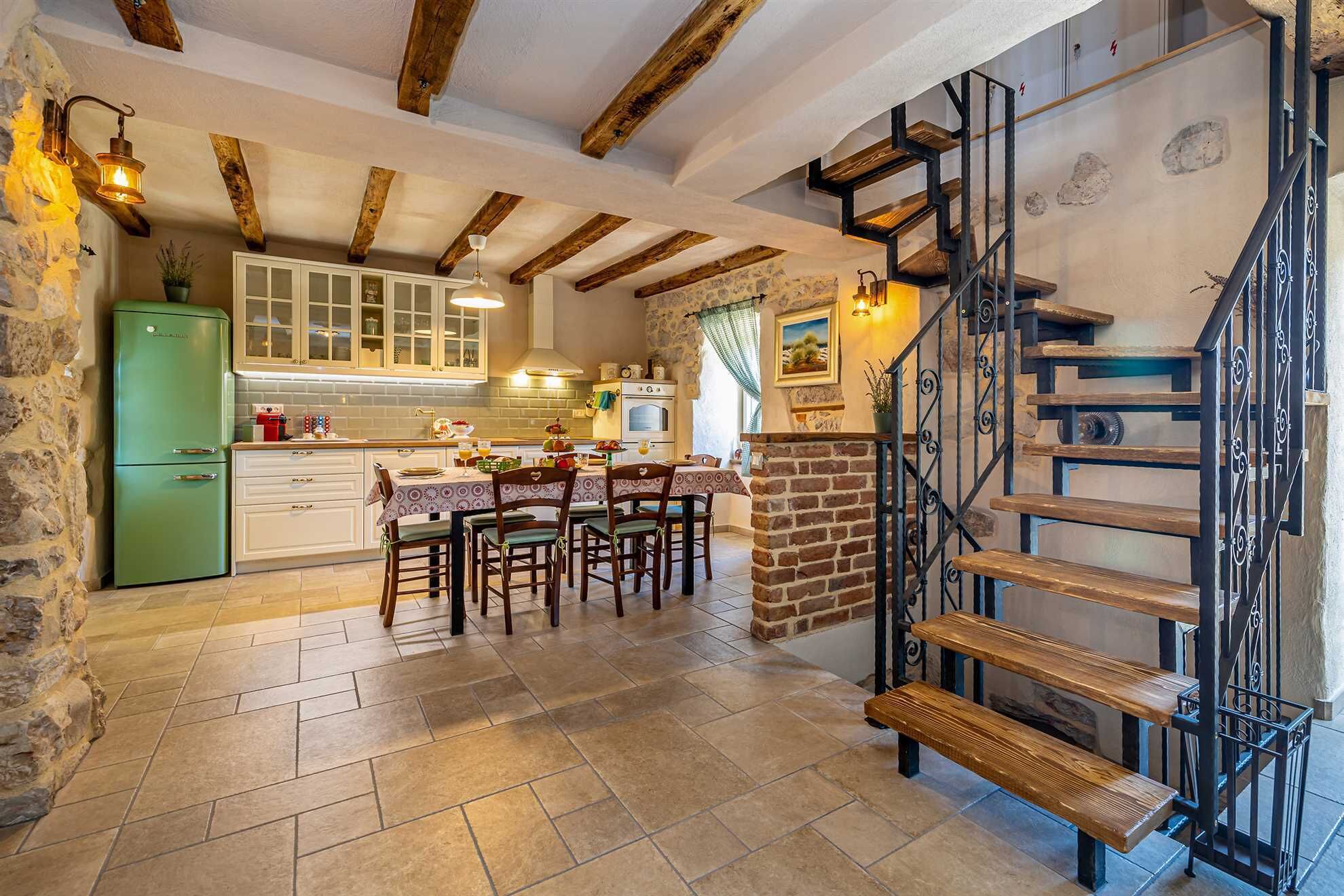 A rustic kitchen with exposed wooden beams, a green refrigerator, and a dining area with a brick wall and metal staircase.