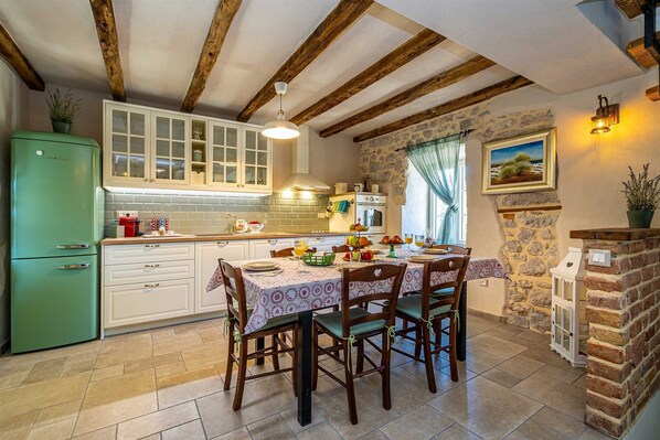 A kitchen with a dining area, featuring exposed wooden beams, a green refrigerator, and a stone wall.