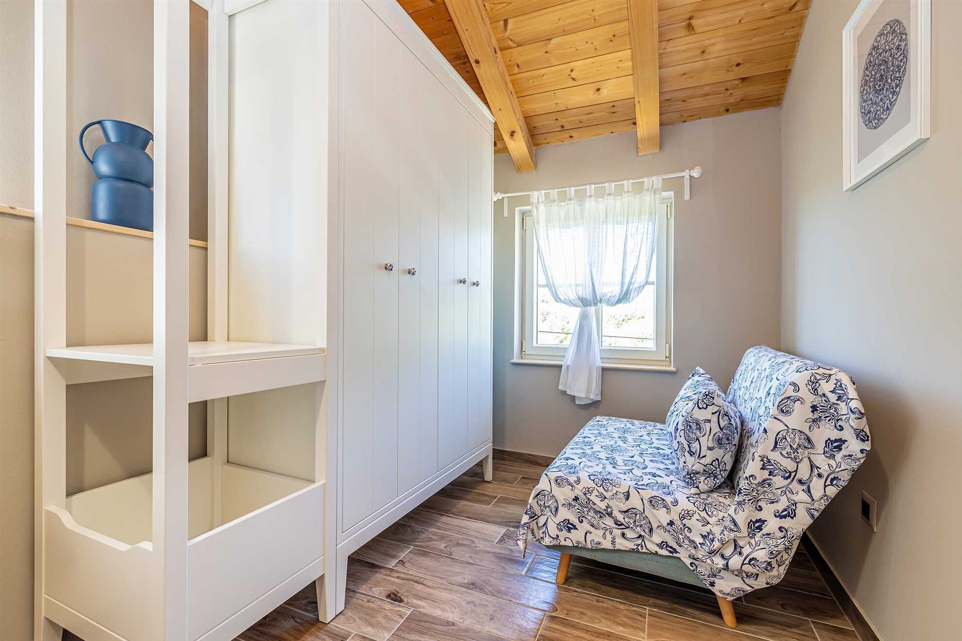Ein Zimmer mit Einbauschrank, einem Fenster mit durchscheinenden Vorhängen, einem gemusterten Sofa und einer Holzbalkendecke.