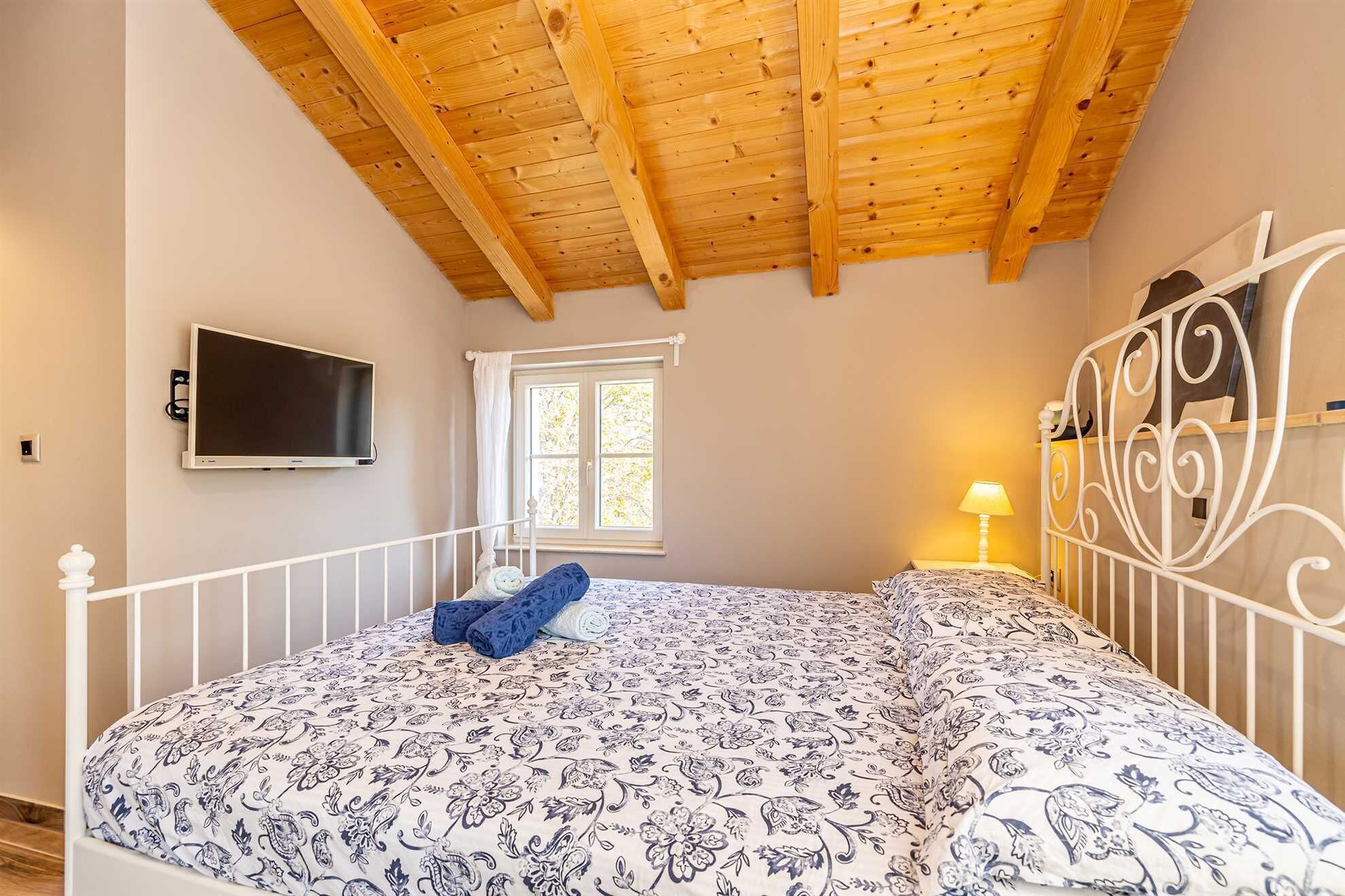 A bedroom with a wooden ceiling, a bed with patterned bedding, a TV mounted on the wall, and a window with curtains.