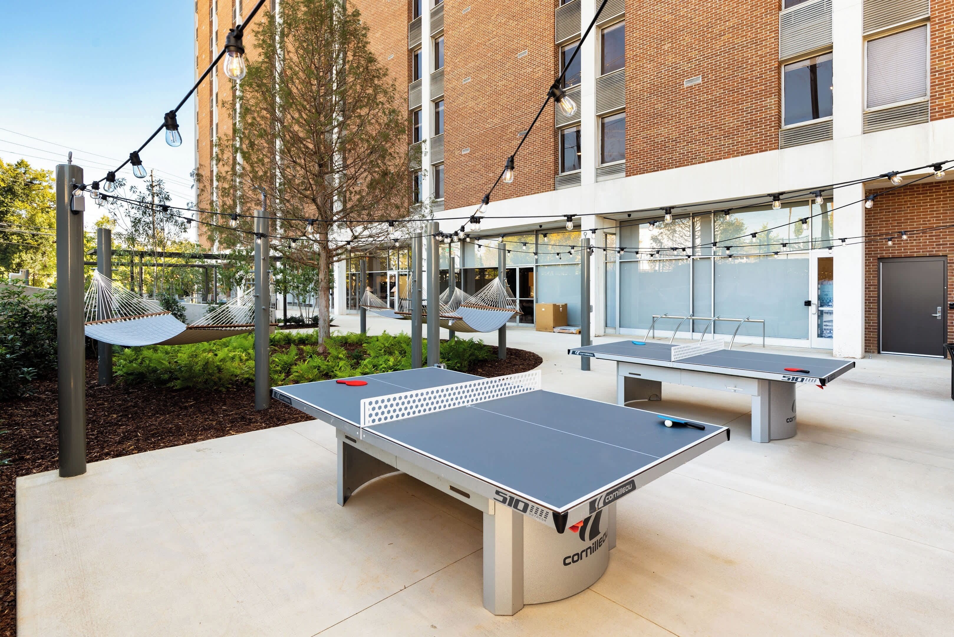 Una cancha de tenis de mesa al aire libre con mesas y redes, rodeada por un edificio con grandes ventanales y fachada de ladrillo.