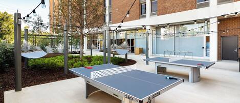 Una cancha de tenis de mesa al aire libre con mesas y redes, rodeada por un edificio con grandes ventanales y fachada de ladrillo.