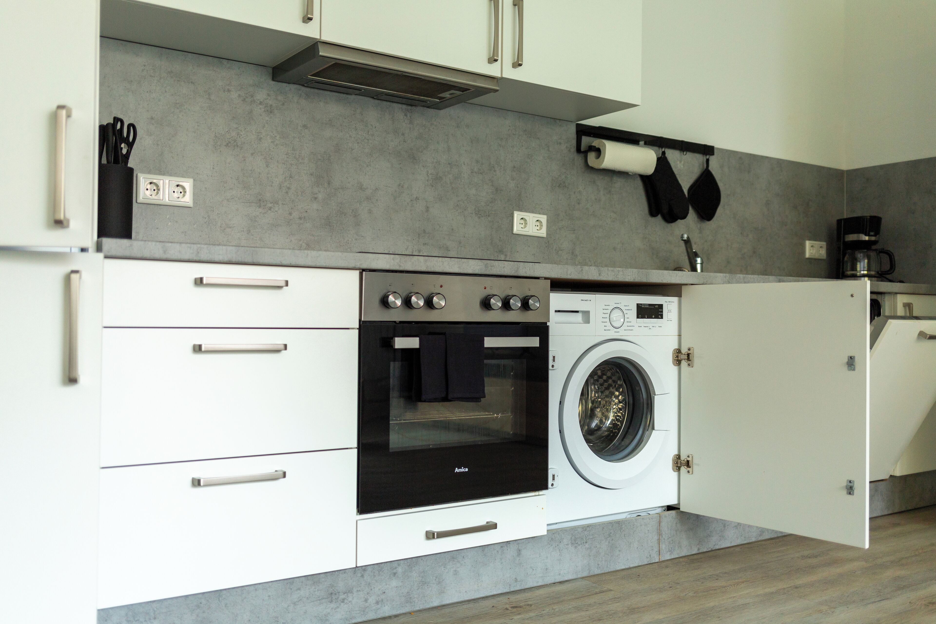 Une cuisine compacte avec des meubles blancs, un four encastré, un lave-linge et une machine à café.
