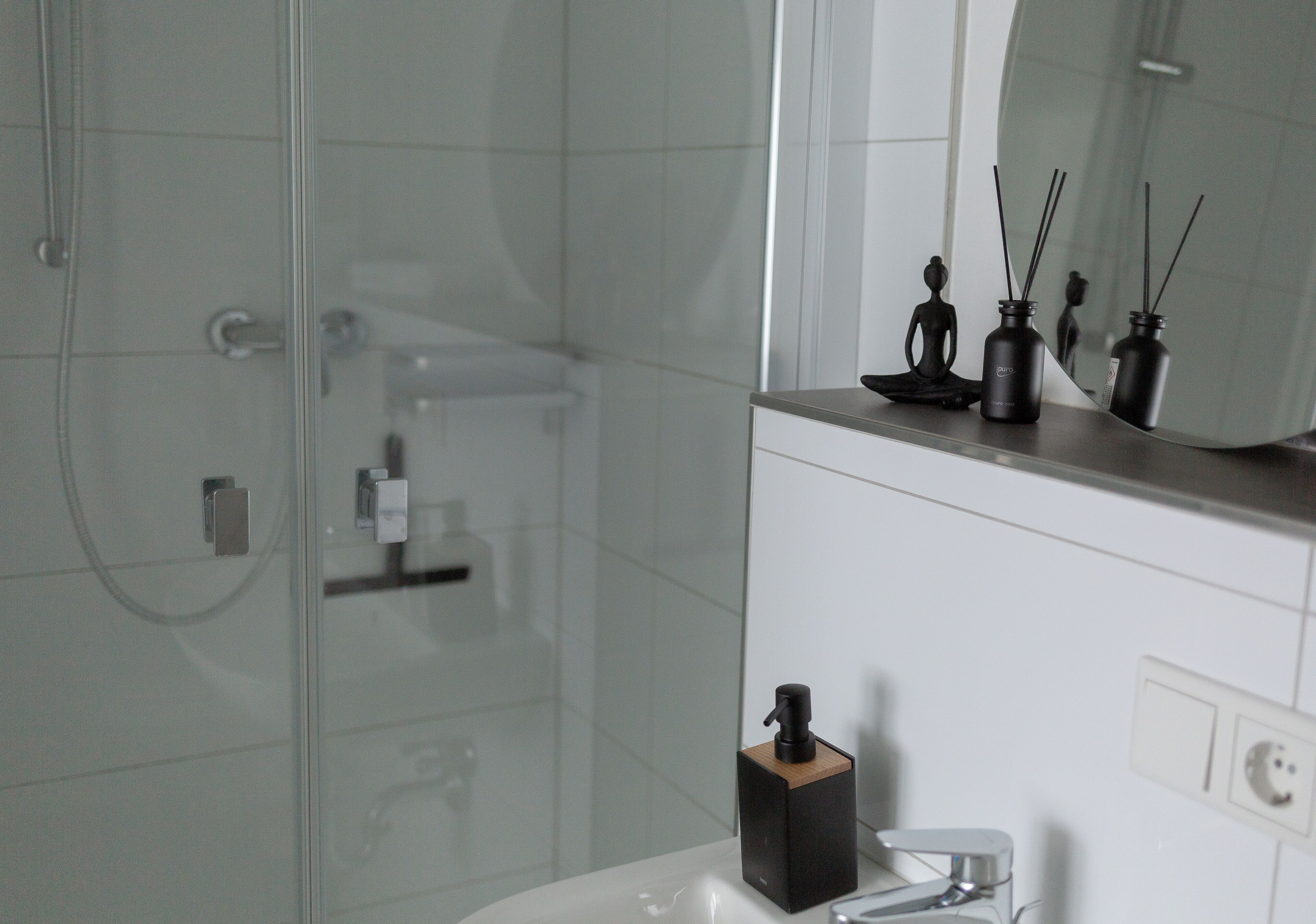 A modern bathroom with a glass shower enclosure, a white sink, and a mirror above it. There are two black reed diffuser candles on the sink's edge.