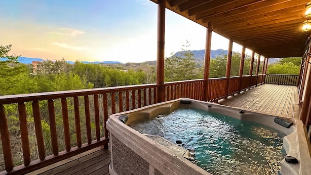 Una terraza de madera con un jacuzzi, barandas de madera y vistas a montañas y vegetación.