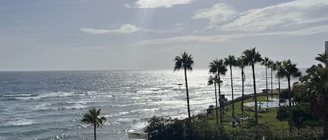 A coastal view with a beach, palm trees, and a building with a thatched roof.