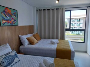 Une chambre d’hôtel avec deux lits, une grande fenêtre donnant sur des immeubles et un tableau coloré représentant des poissons accroché au mur.