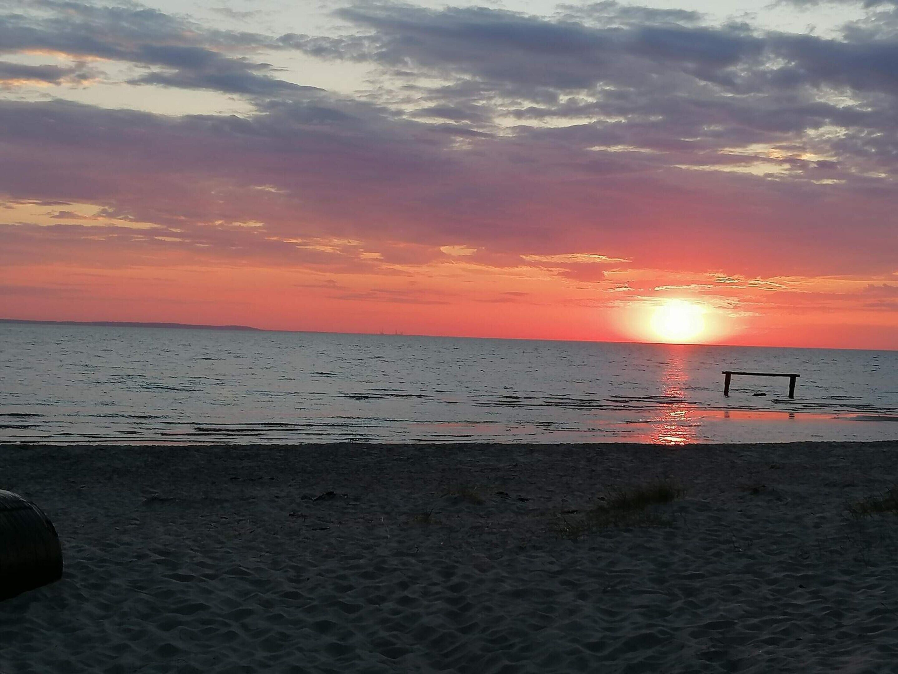 Una playa con un atardecer, mar tranquilo y un banco.